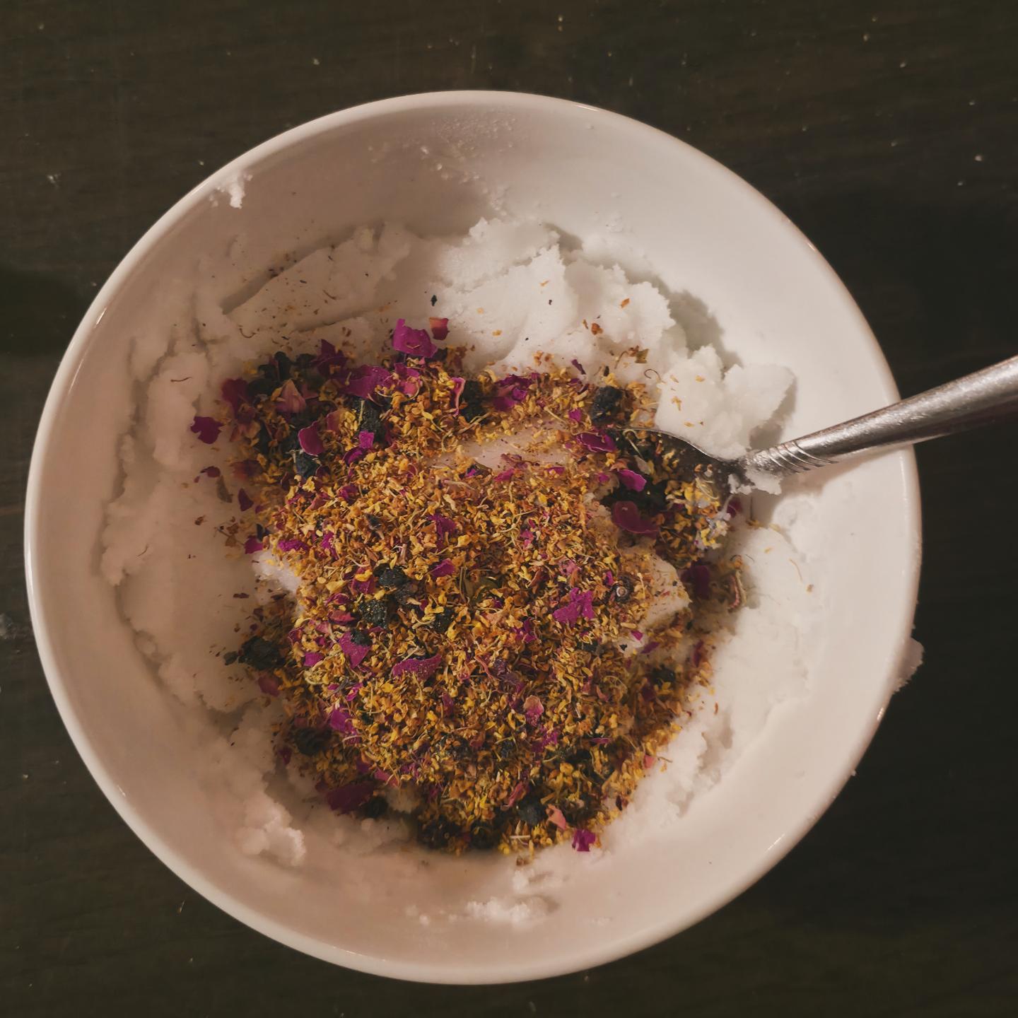 White bowl of salt scrub topped with dried flowers and herbs